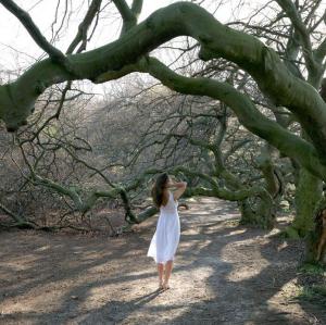 A barefoot woman in a white dress stands, her back toward us, on dirt under a big, gnarled tree branch.