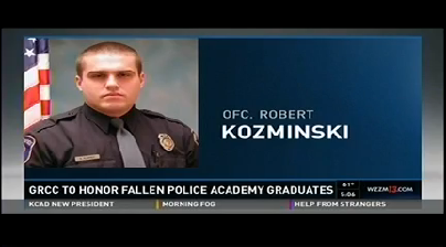 A television screen shows a photo of a police officer: GRCC to honor fallen Police Academy graduates. Ofc. Robert Kozminski.